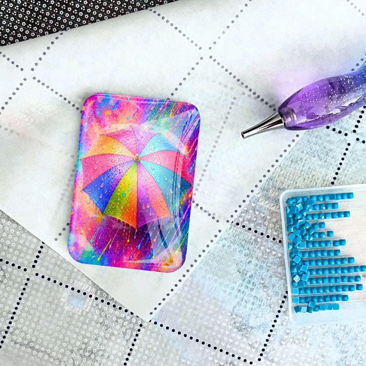 Diamond Painting Cover Minder aus Glas mit buntem Regenschirm auf Leinwand neben Stift und Steinchen.