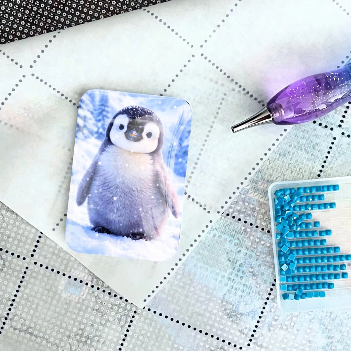 Diamond Painting Zubehör: Cover Minder Winterpieps mit Pinguin auf Leinwand neben Glitzerstift und Steinchen.